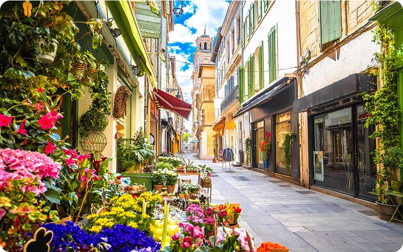 A charming European street with colorful flower shops, traditional buildings with green shutters, and a peaceful cobblestone walkway lined with potted plants and awnings